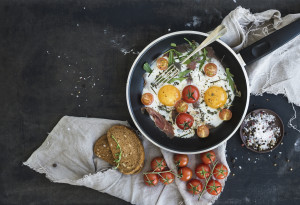 Pan of fried eggs, bacon and cherry-tomatoes with bread on dark table surface, top view